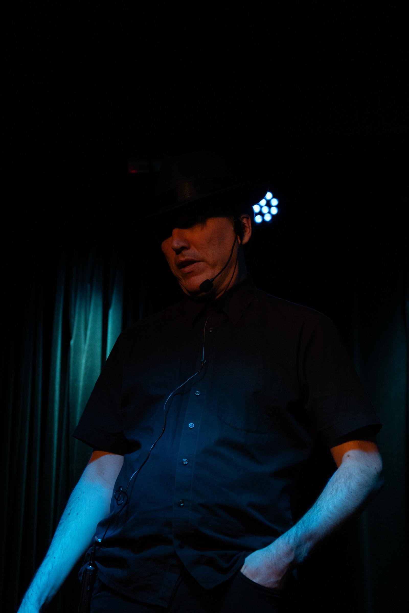 Solo performer in a fedora and headset under dim stage light