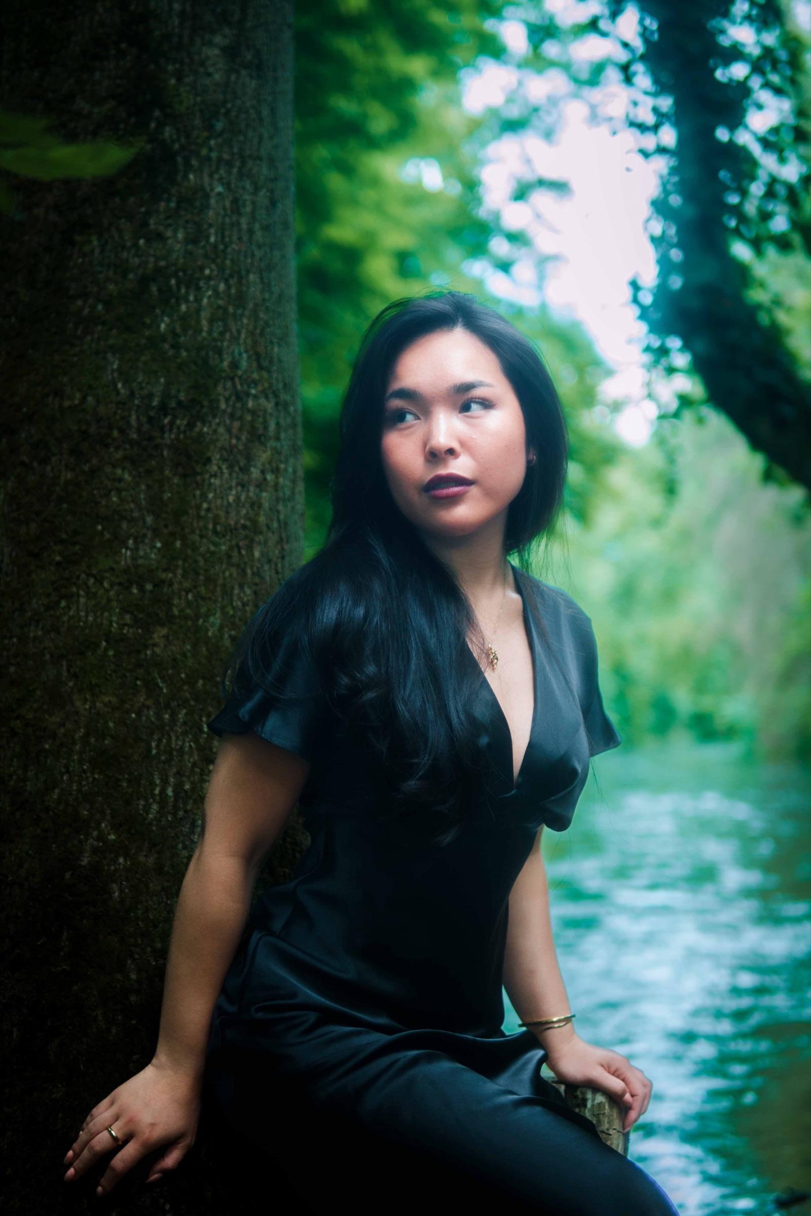Woman in black leaning against a mossy trunk by a river, glancing aside
