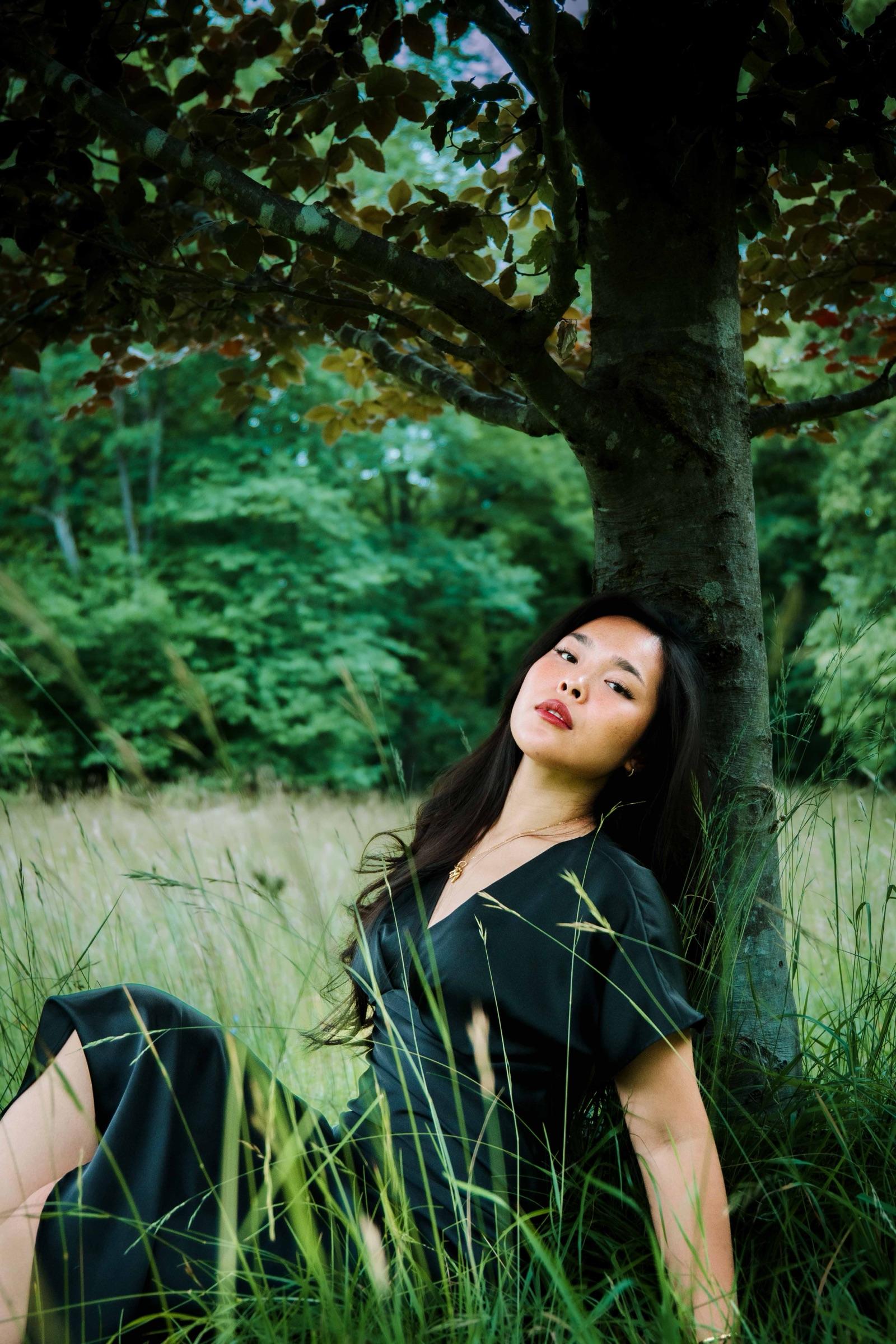 Woman in a black dress resting against a tree trunk in tall grass